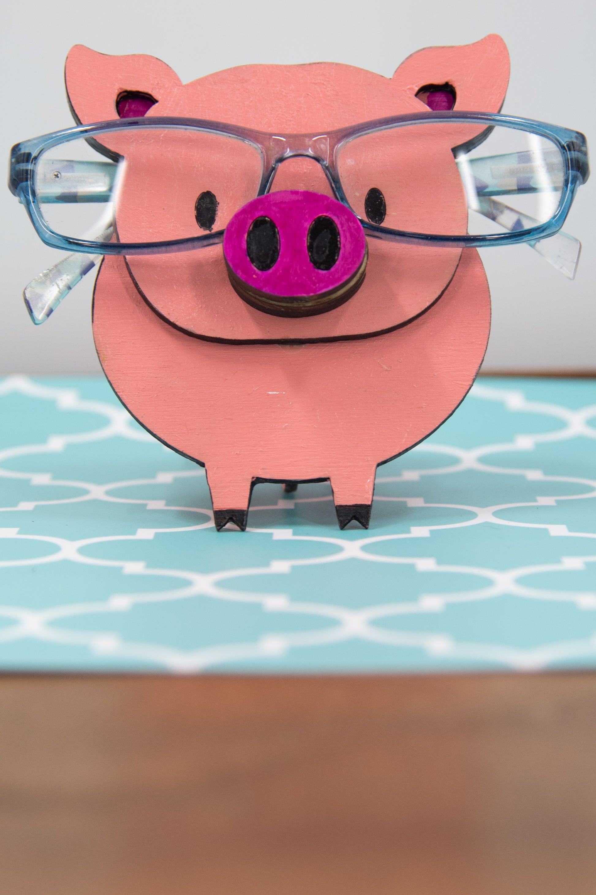 Handcrafted wooden pig-shaped eyeglass stand with pink finish and black details, holding glasses on its snout, ideal for desks or gift giving.